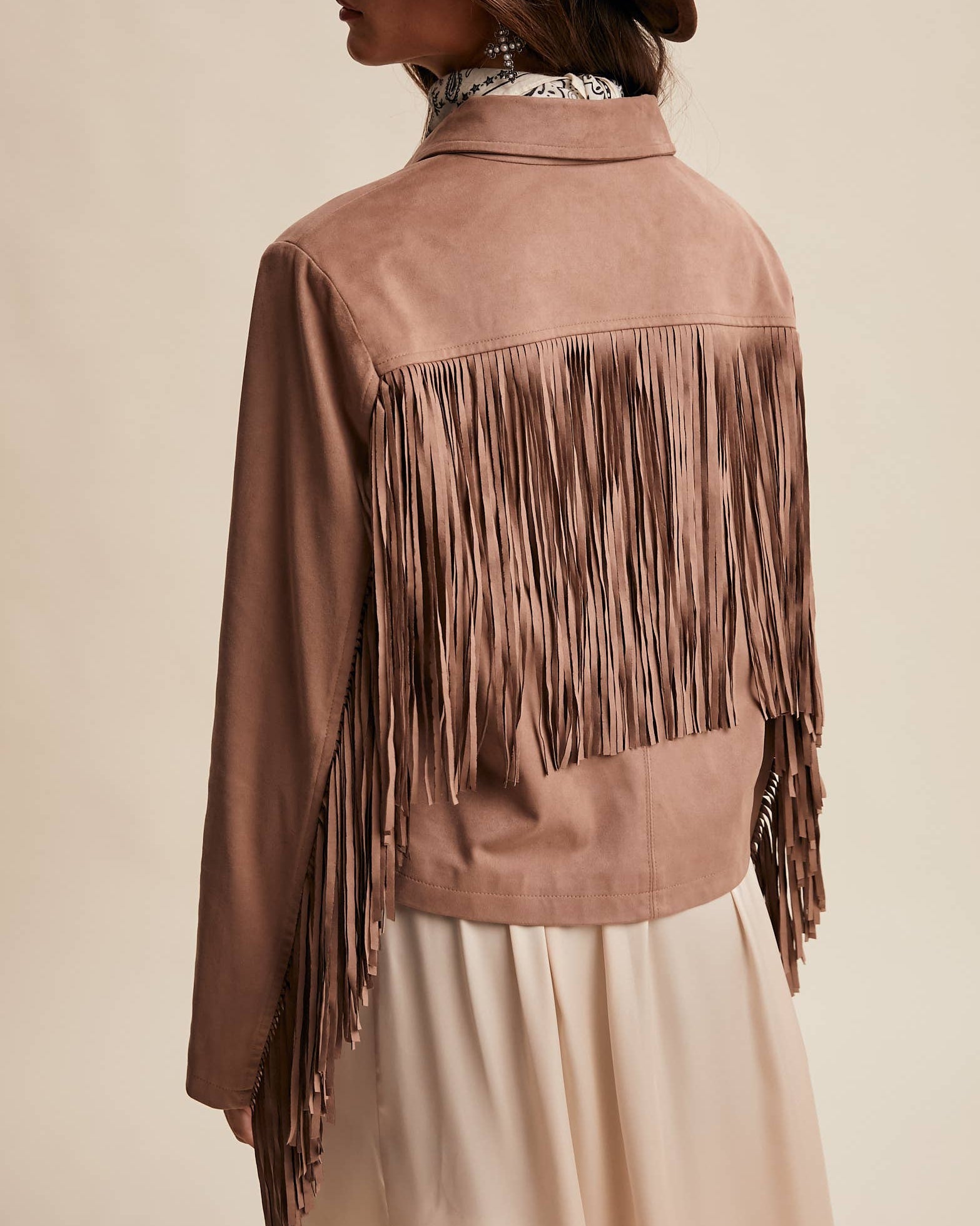Person wearing a brown suede jacket with fringe and a matching hat on a beige background