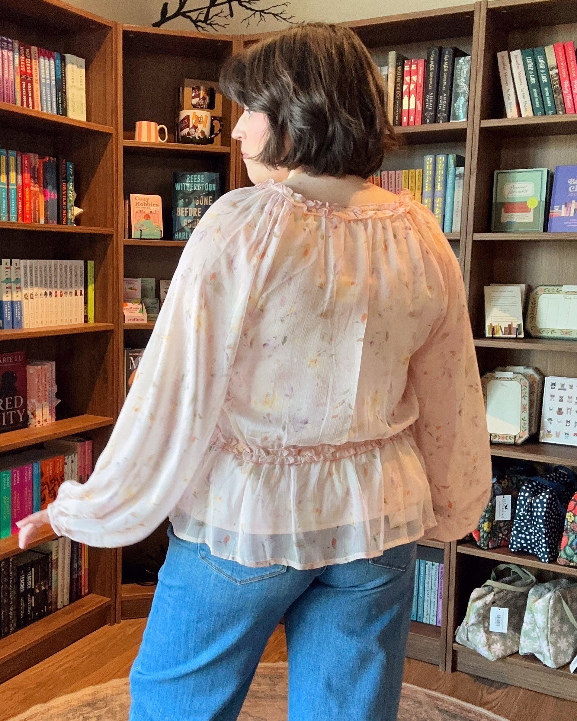 Person wearing a sheer blouse and blue jeans standing in front of bookshelves.