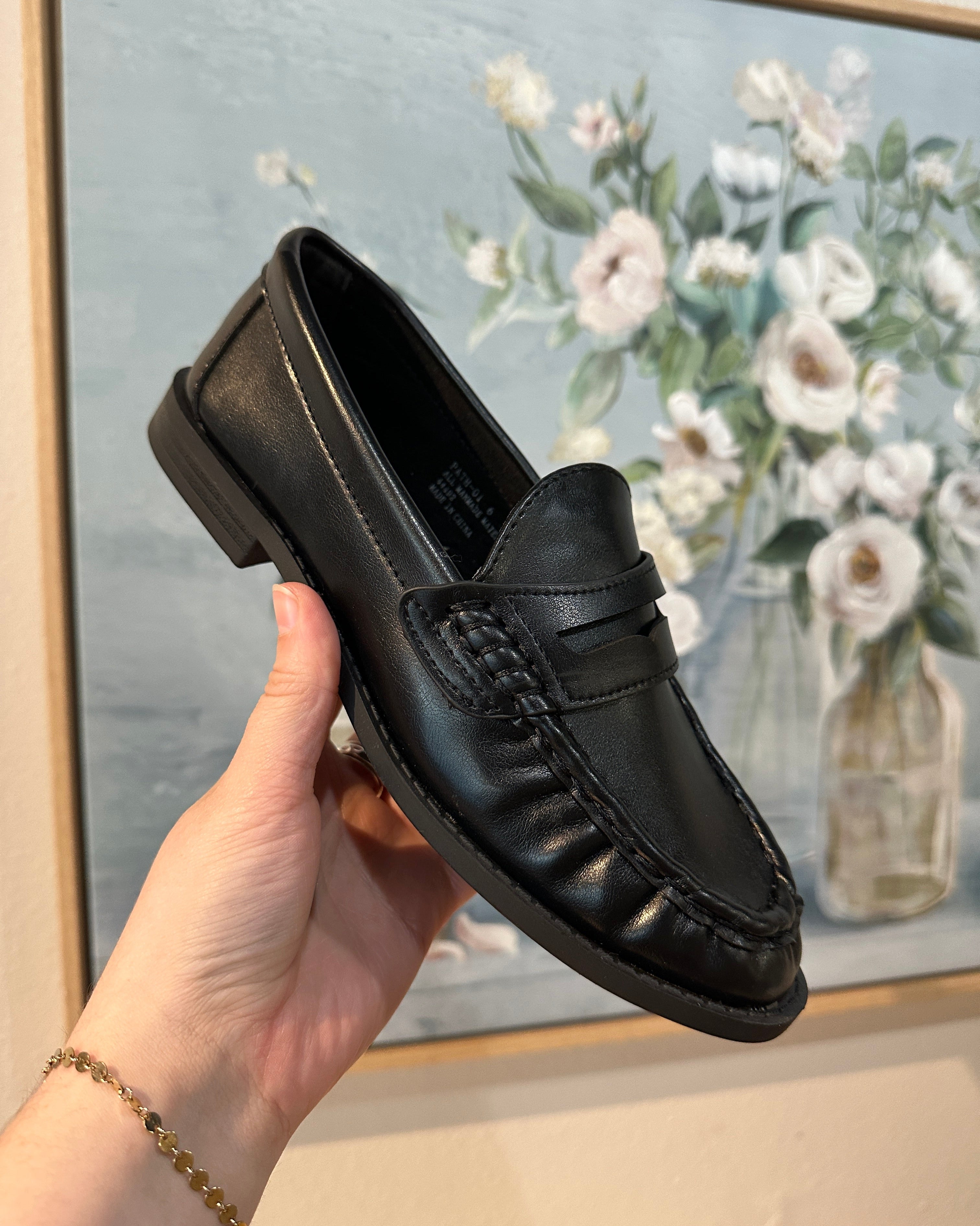 Black loafer shoe held by a hand with a floral painting in the background