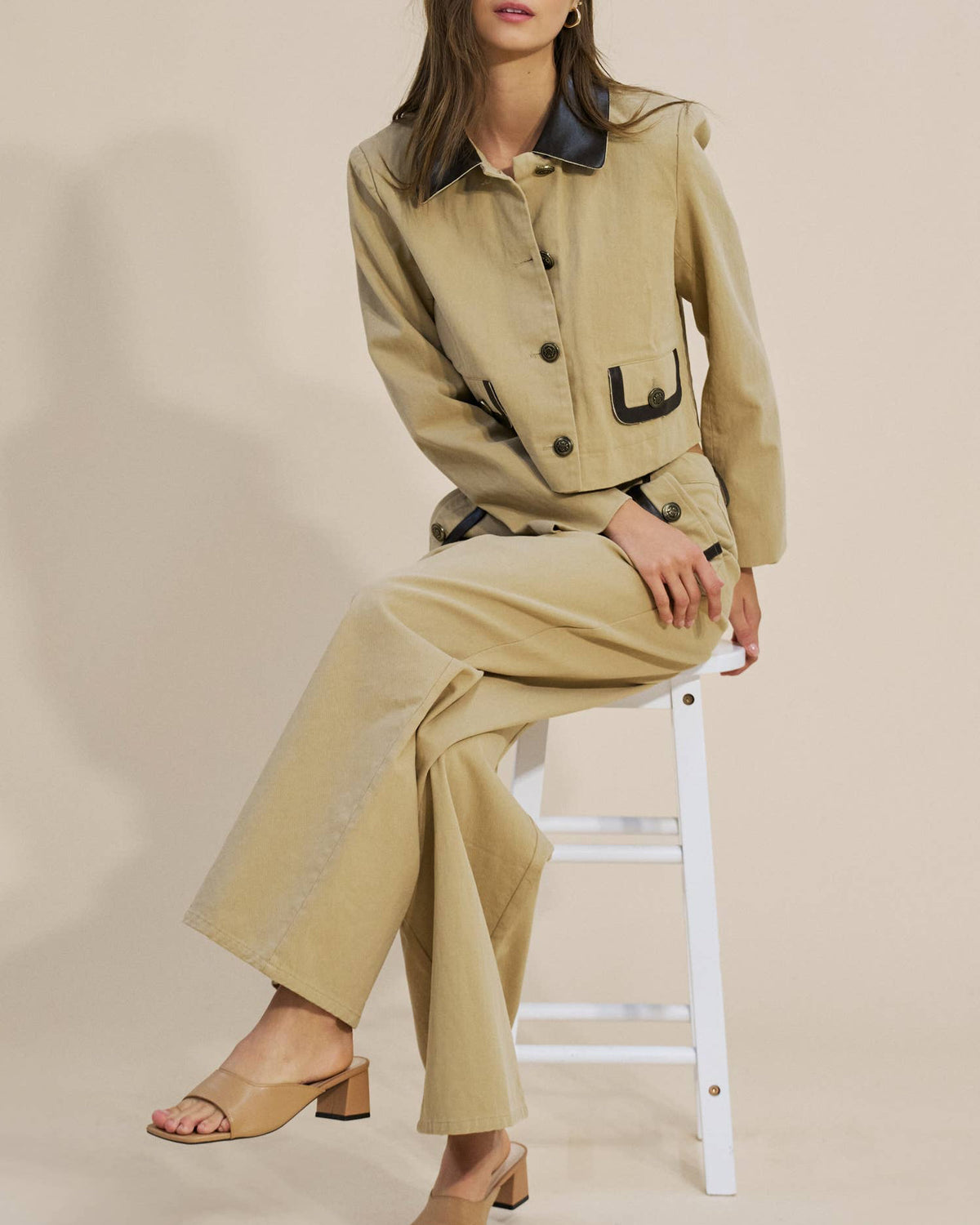 A woman models a beige two-piece linen set consisting of a cropped button-up jacket with dark contrast collar and pocket trim, paired with matching wide-leg trousers, while sitting on a white stool.