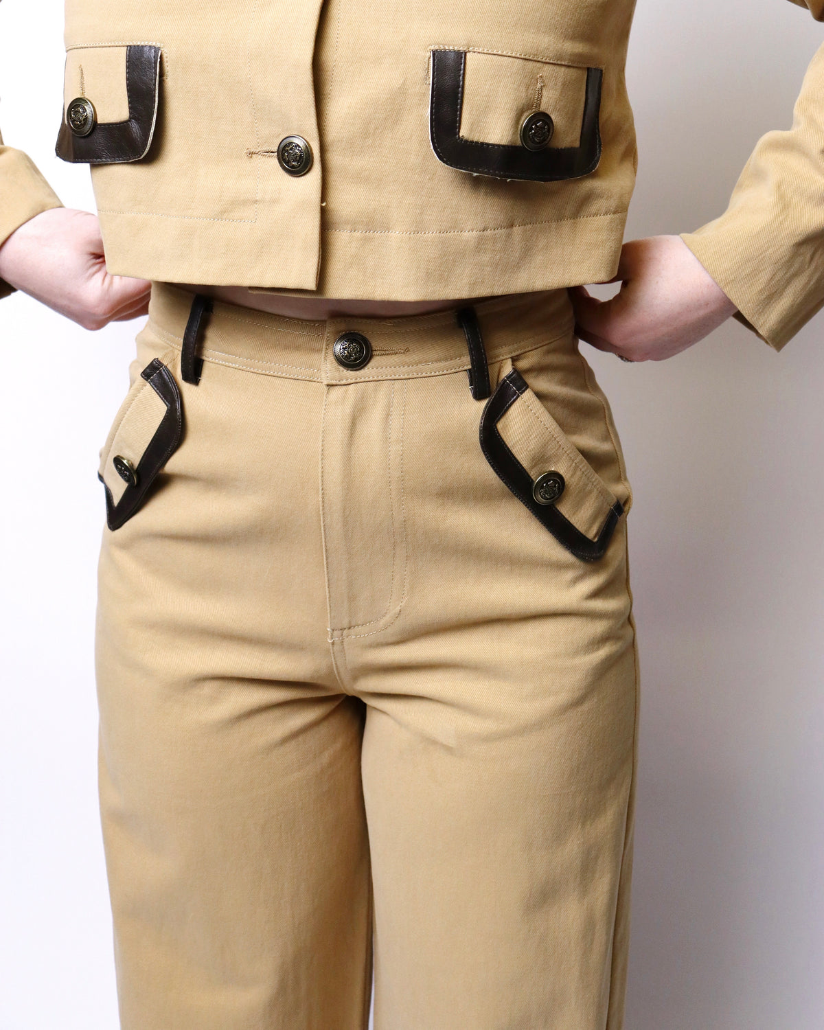 A close-up of a person wearing tan utility-style pants and a matching cropped jacket with black contrast trim and flap pockets, standing against a plain light background.