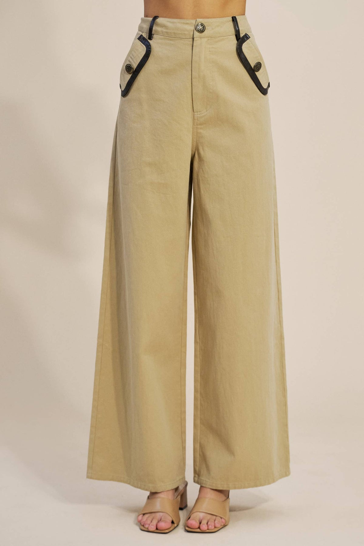 A person wearing high-waisted, wide-leg beige pants with black contrast trim along the pocket edges and front button closure, paired with clear high-heel sandals against a plain beige studio background.