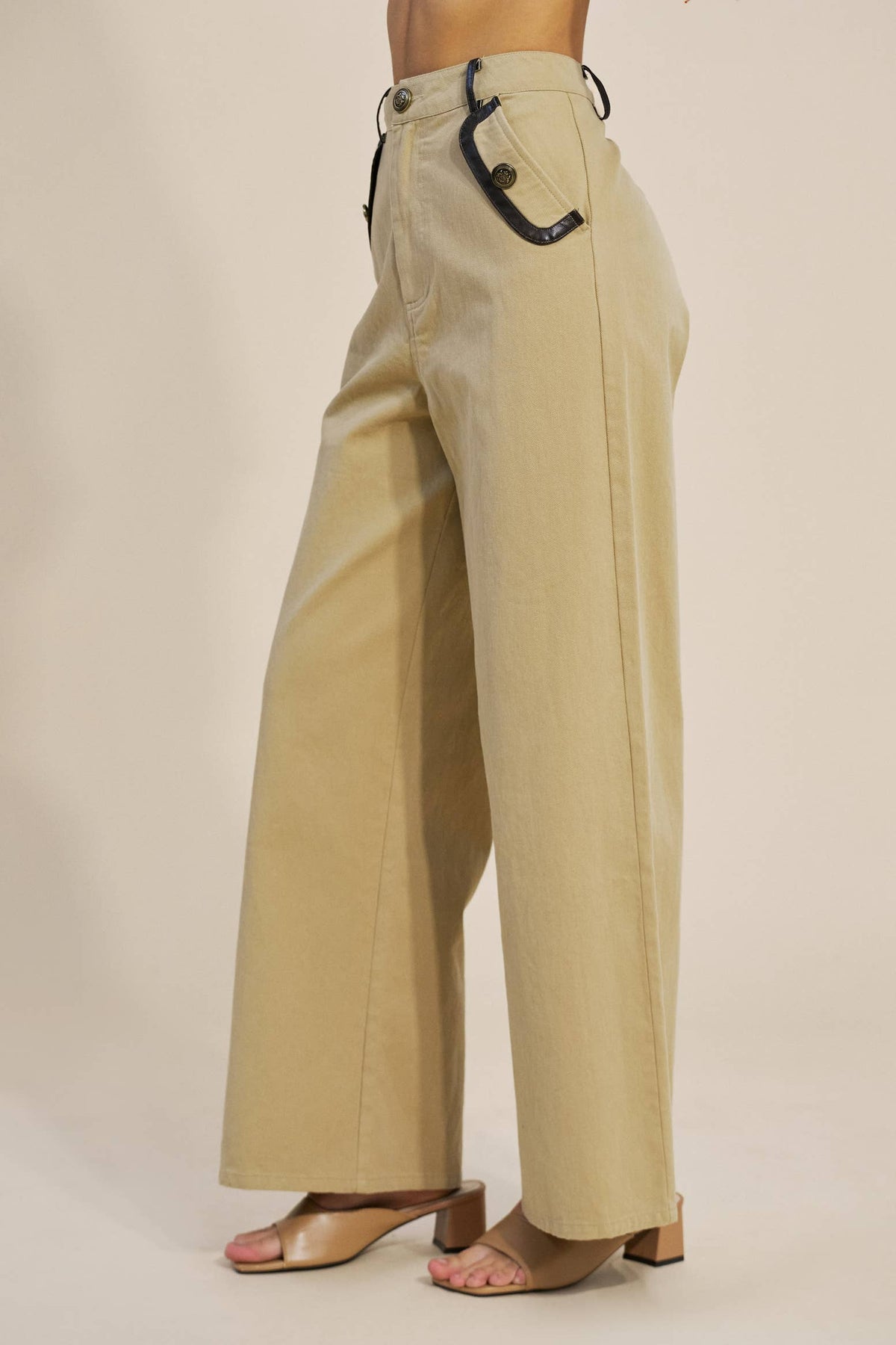 A person wearing high-waisted, wide-leg beige pants with black leather accents on the waistband and side pocket, paired with tan heeled sandals against a plain beige studio background.