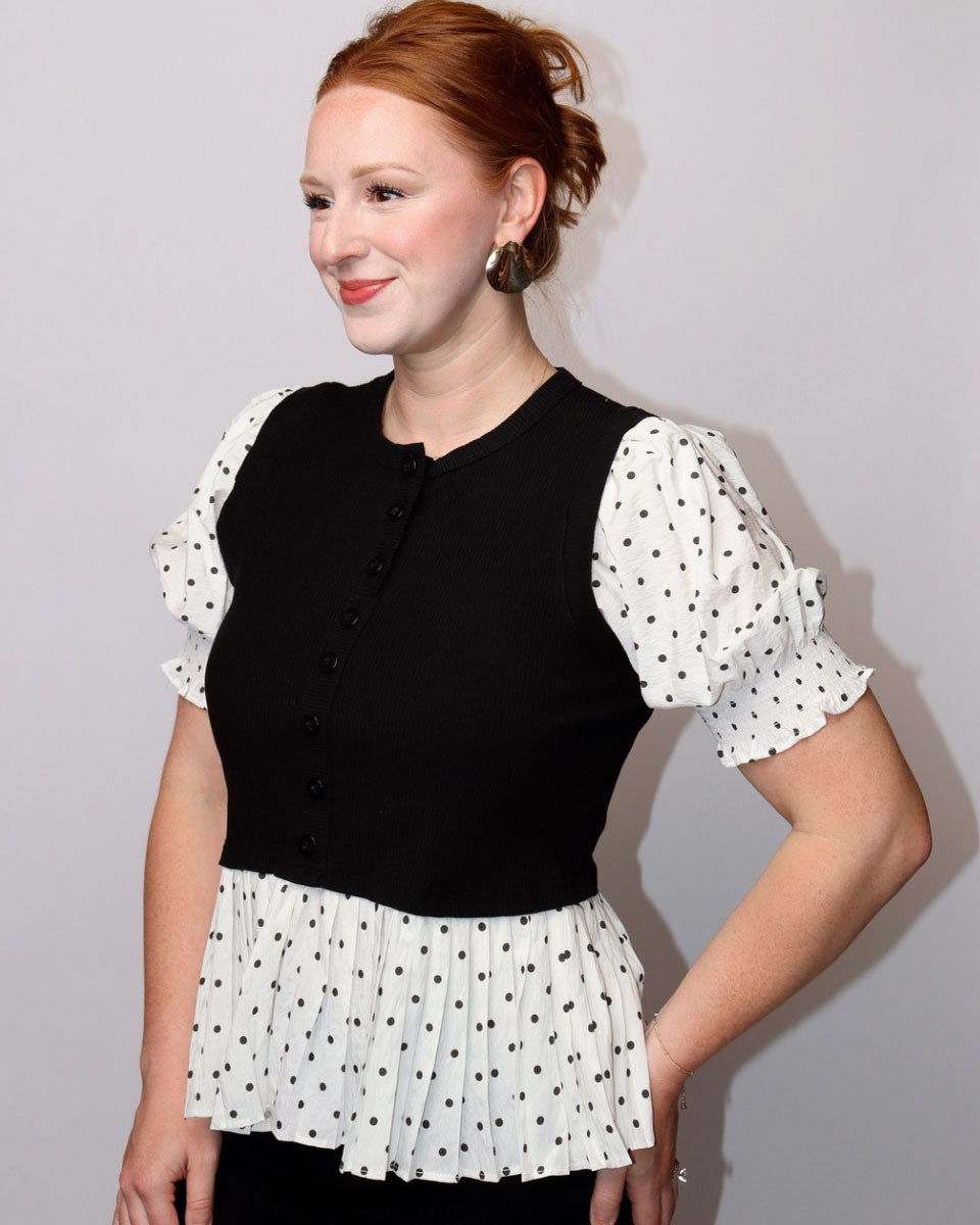A woman with red hair in an updo wearing a black vest over a white puff-sleeve top with black polka dots and a matching polka-dot peplum hem, posed against a plain light gray background.