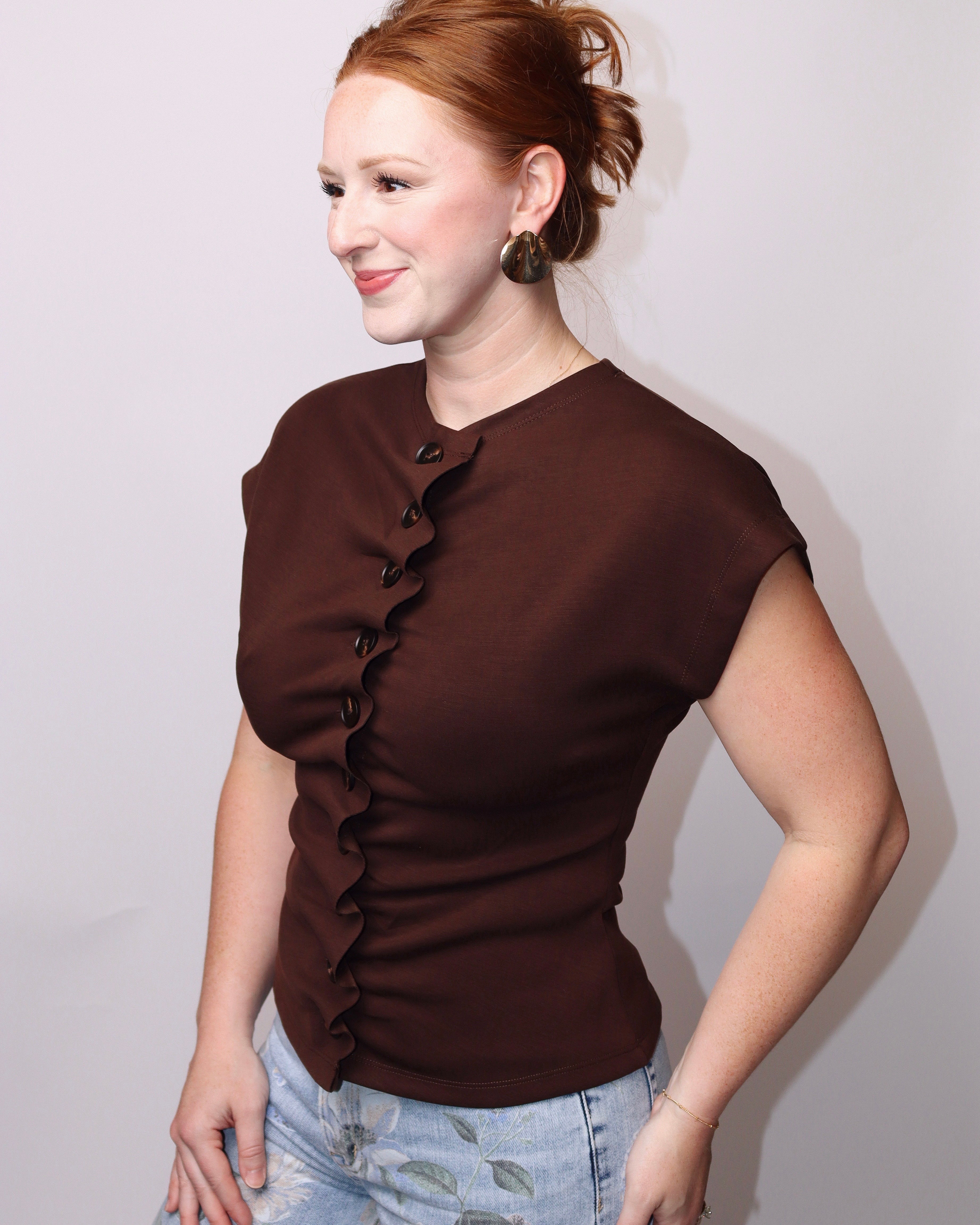 A woman with red hair in a bun wearing a dark brown ruffled short-sleeve top and light blue jeans, standing against a plain light gray background.
