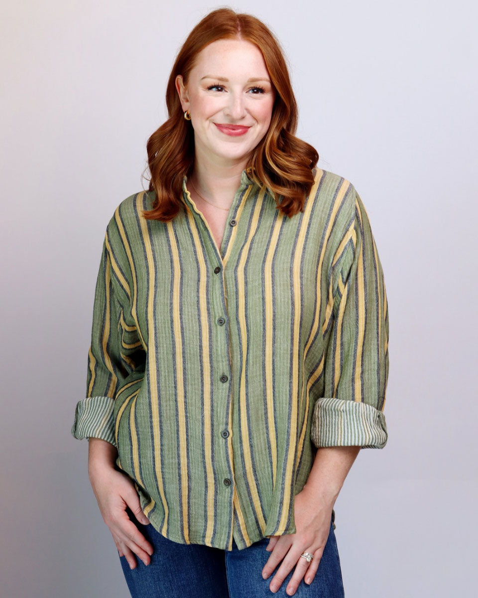 A smiling woman with red hair wearing a green and yellow vertically striped button-down shirt with rolled-up sleeves, paired with blue jeans, standing against a light gray studio background.