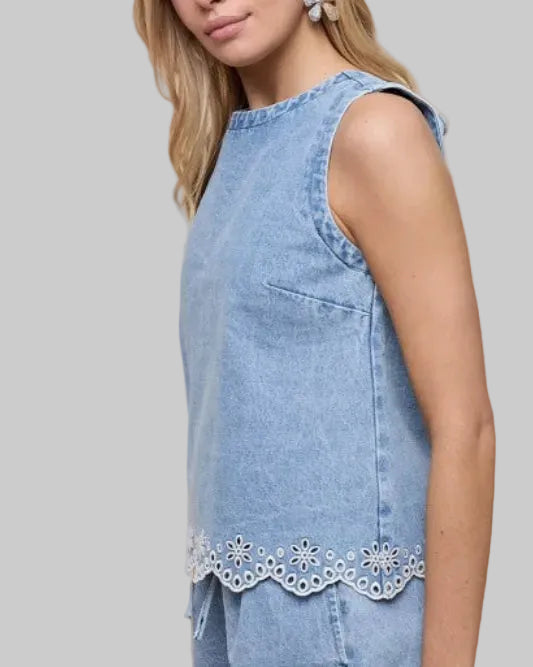 Model wearing a light blue sleeveless denim with eyelet embroidery at hem top on a neutral background.  Side view