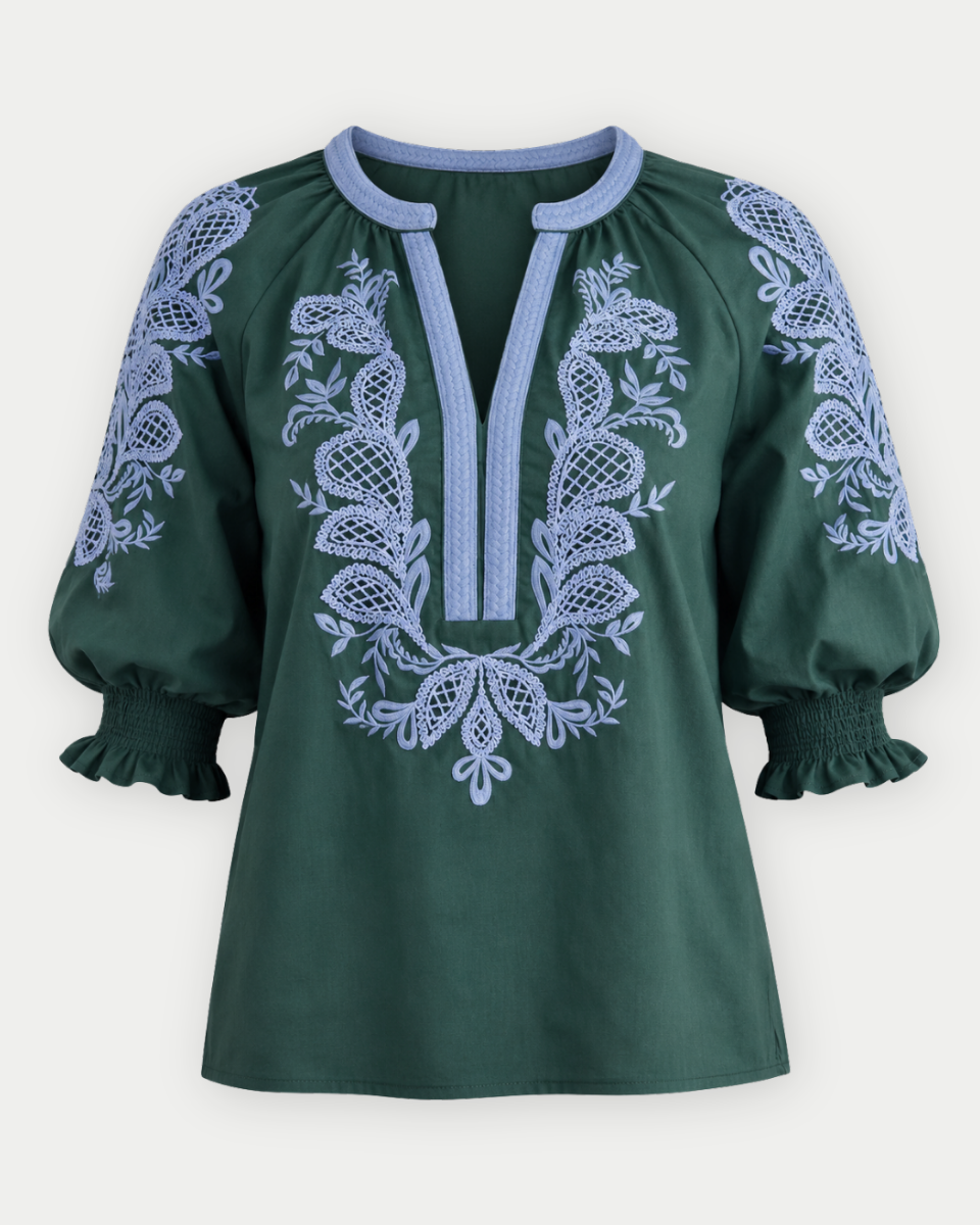 A dark green blouse with light blue embroidery and trim, featuring a split neckline, puff sleeves, and a smocked hem, shown against a light gray background.