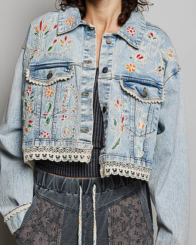 Model wearing a denim jacket with embroidery and beading.  Greens, oranges, and reds in the embroidery and beading.  Lace accents at the waist and cuff.