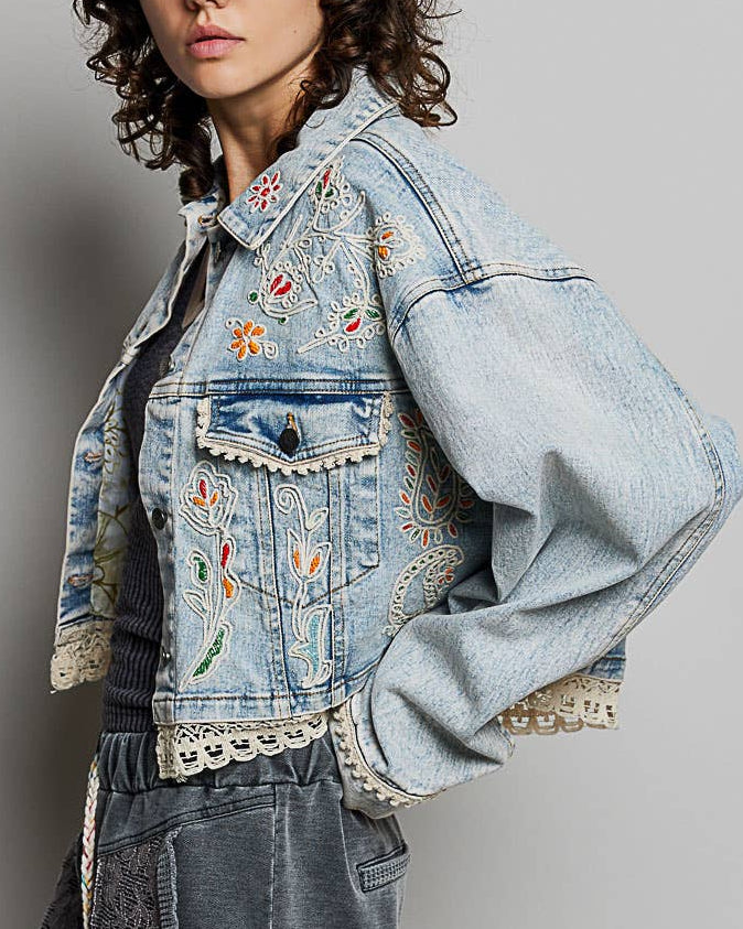 Model wearing a denim jacket with embroidery and beading.  Greens, oranges, and reds in the embroidery and beading.  Lace accents at the waist and cuff.