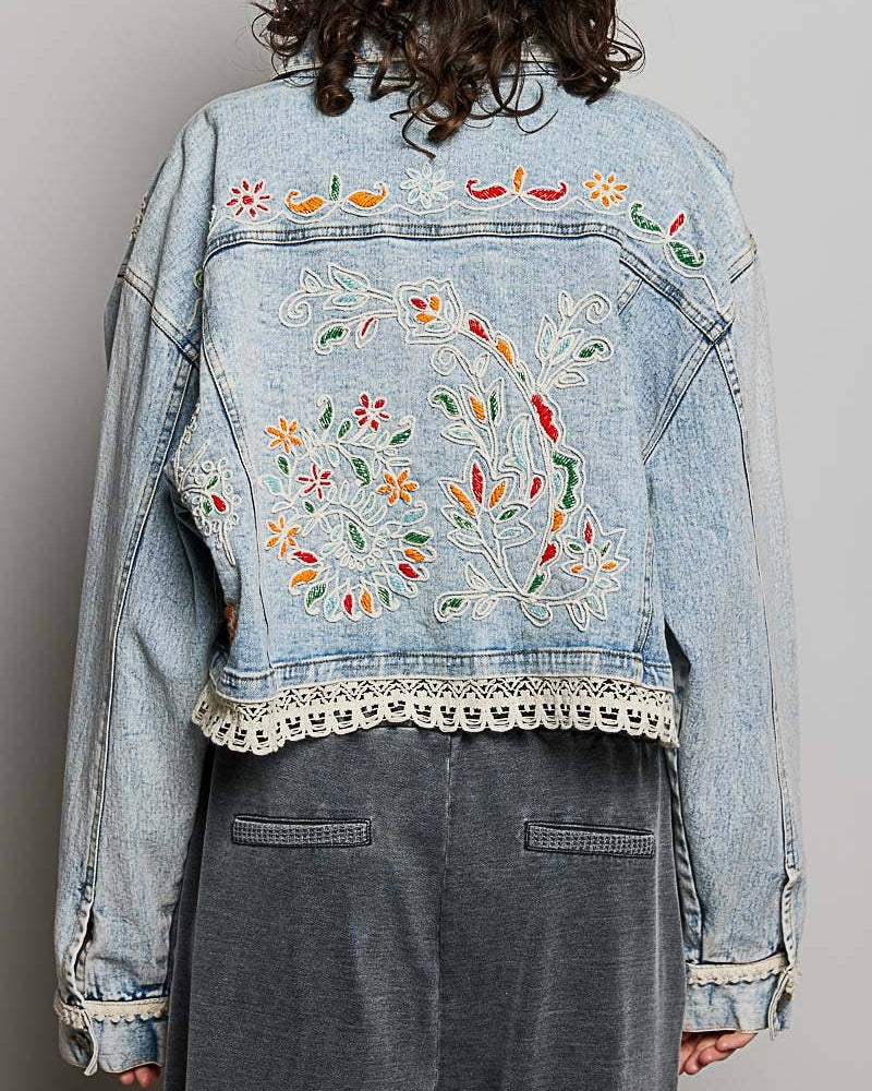 Model wearing a denim jacket with embroidery and beading.  Greens, oranges, and reds in the embroidery and beading.  Lace accents at the waist and cuff.