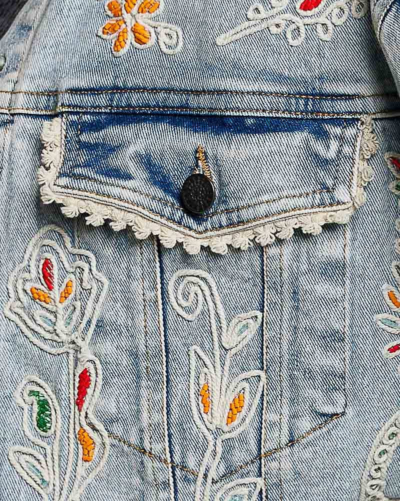 Model wearing a denim jacket with embroidery and beading.  Greens, oranges, and reds in the embroidery and beading.  Lace accents at the waist and cuff.