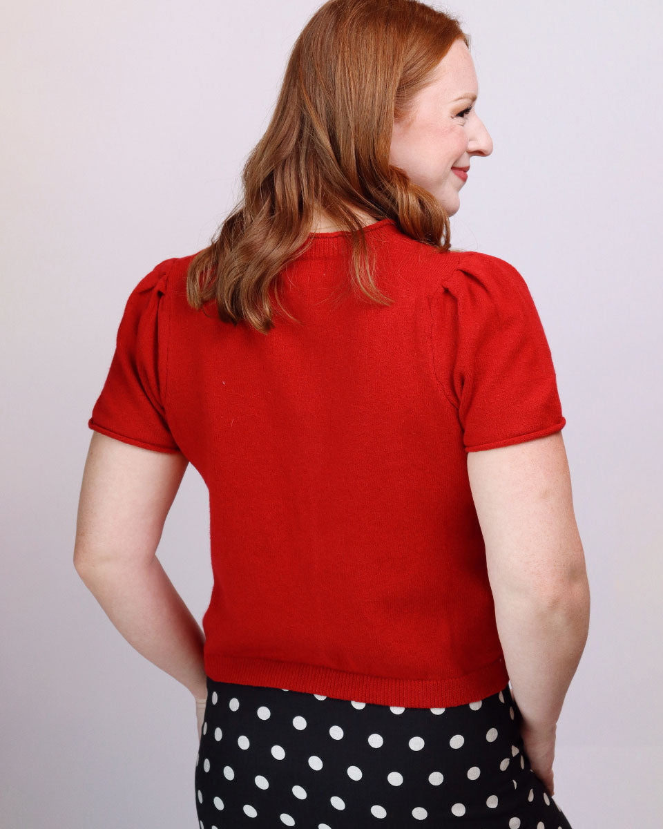 A woman with red hair is shown from the back wearing a bright red short-sleeve knit top and a black skirt with white polka dots, set against a plain light gray background.