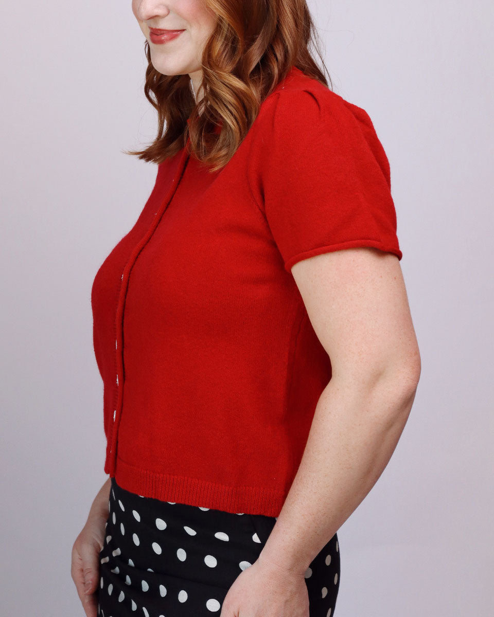 A woman wearing a bright red short-sleeve knit cardigan with a button front, paired with a black and white polka dot skirt, shown from a side profile against a light gray studio background.