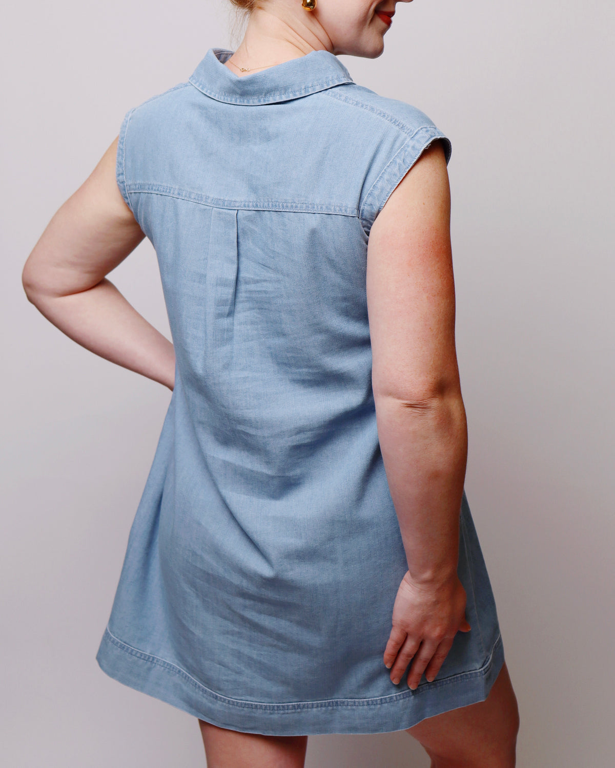 A woman wearing a light blue sleeveless denim dress, shown from the back against a plain light gray studio background.
