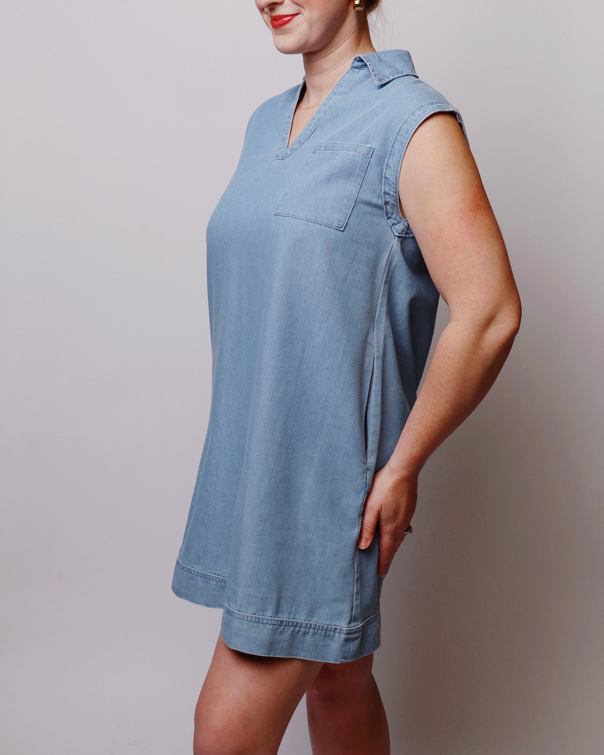 A woman wearing a light blue sleeveless collared mini dress with a chest pocket, shown in a side profile against a plain light gray studio background.