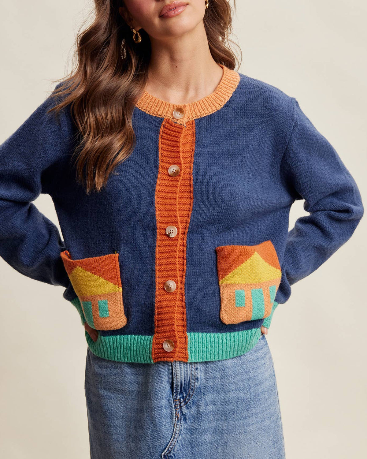 A woman wearing a blue knit cardigan with orange trim, decorative house-shaped pockets, and a turquoise ribbed hem, paired with a denim skirt against a plain beige background.