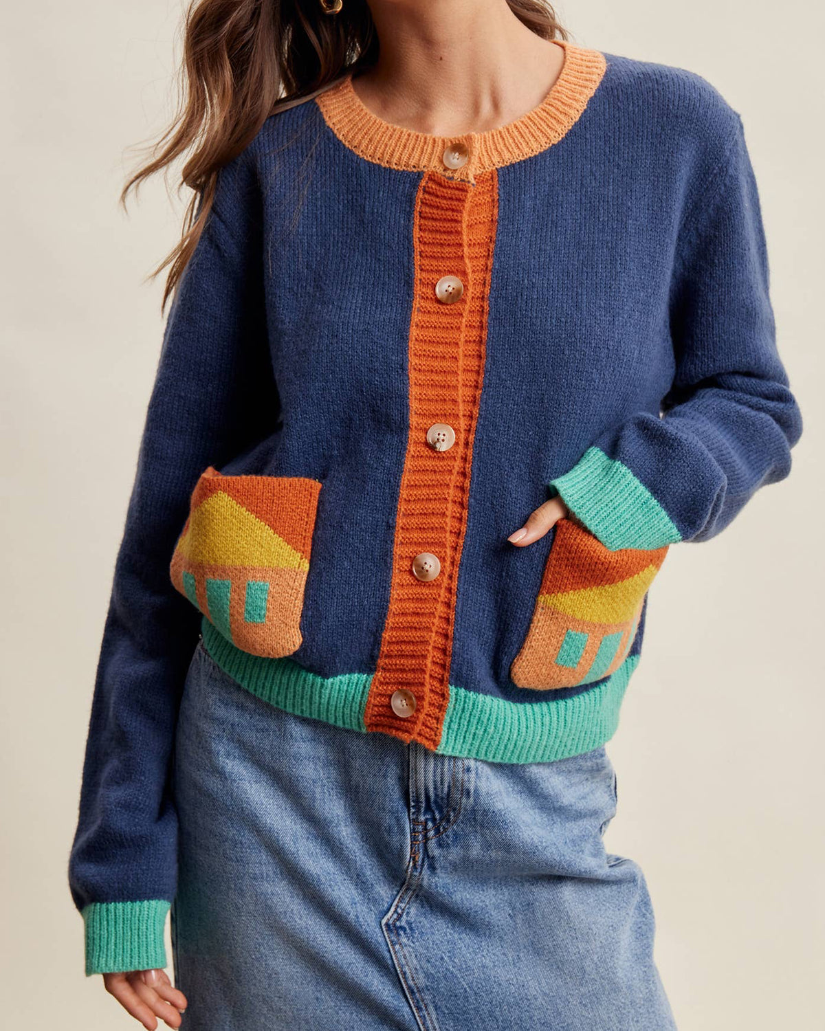 A woman wearing a blue knit cardigan with orange, teal, and yellow geometric patterned details, paired with a denim skirt, against a plain beige background.