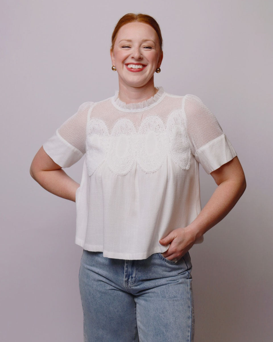 A smiling woman wearing a white short-sleeve blouse with sheer mesh sleeves and a scalloped lace yoke, paired with light blue jeans, against a plain light gray studio background.