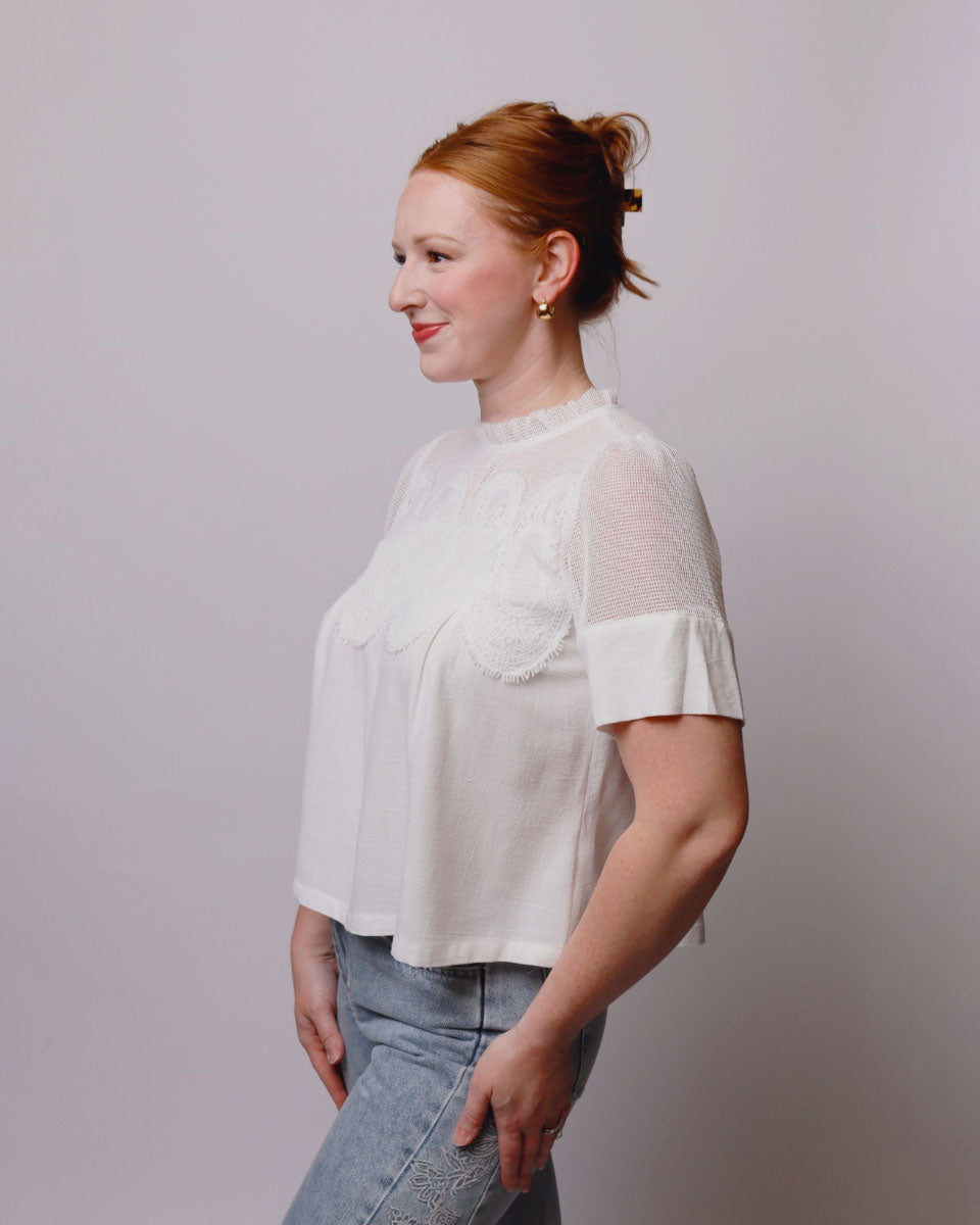 A woman with red hair in a bun wearing a white short-sleeve blouse with sheer mesh sleeves and lace applique on the chest, paired with light blue jeans, shown in profile against a solid light gray background.