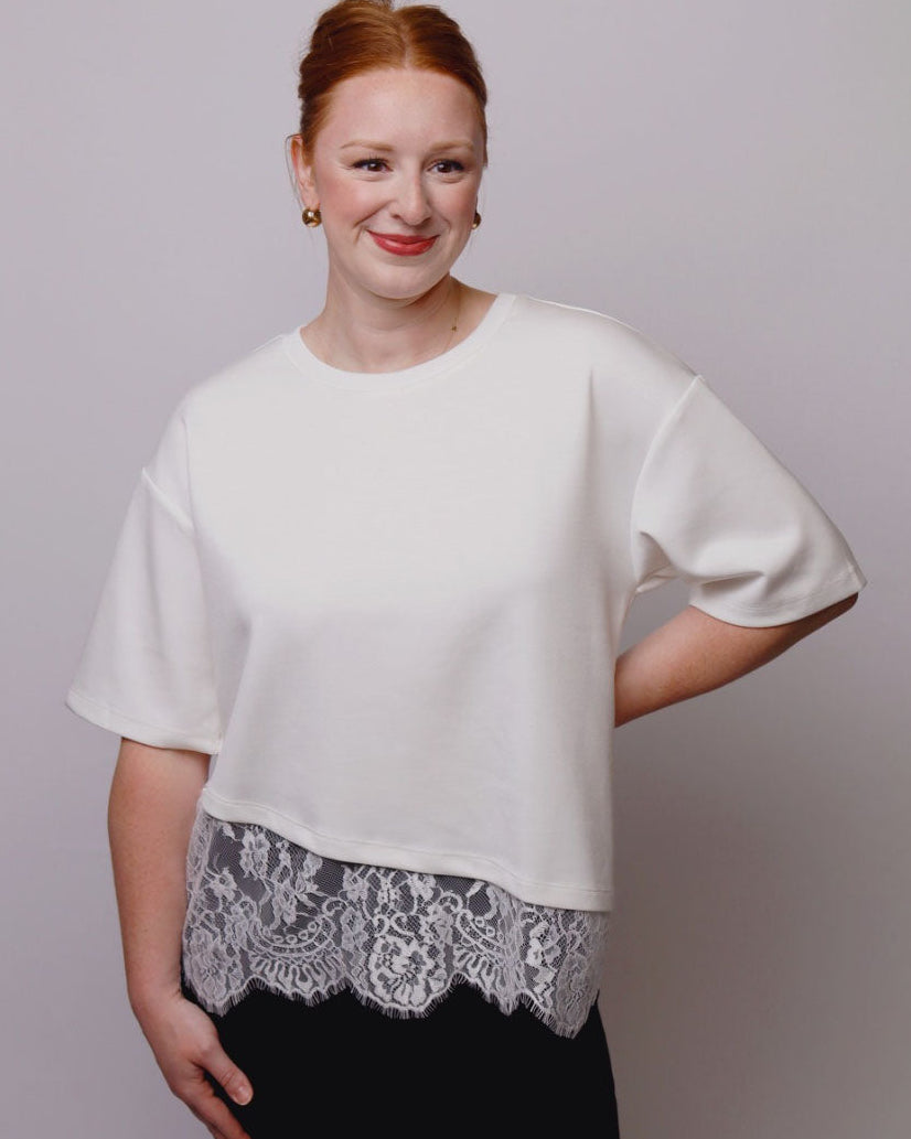 A woman with red hair tied back, wearing a white short-sleeve crop top with a white lace hem detail and black pants, standing against a light gray studio background.