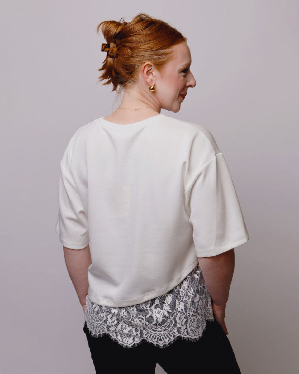 A woman with red hair in a bun is shown from the back wearing an oversized white short-sleeve top, a black and white lace hem detail, and black pants, set against a plain light gray studio background.