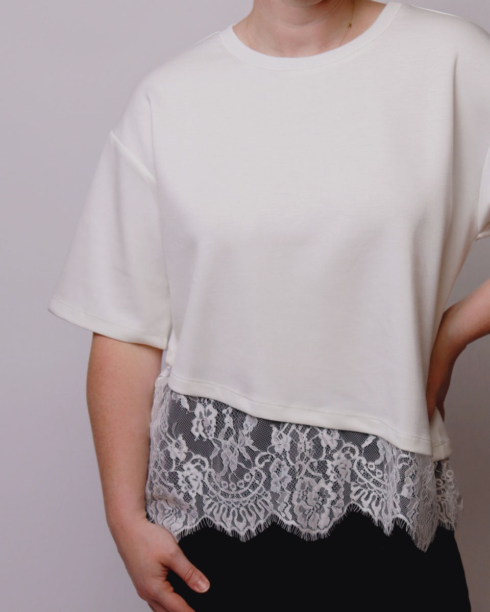 A person wearing a short-sleeve cream-colored top with a sheer white floral lace hem overlay and black bottoms, shown against a plain light gray background.