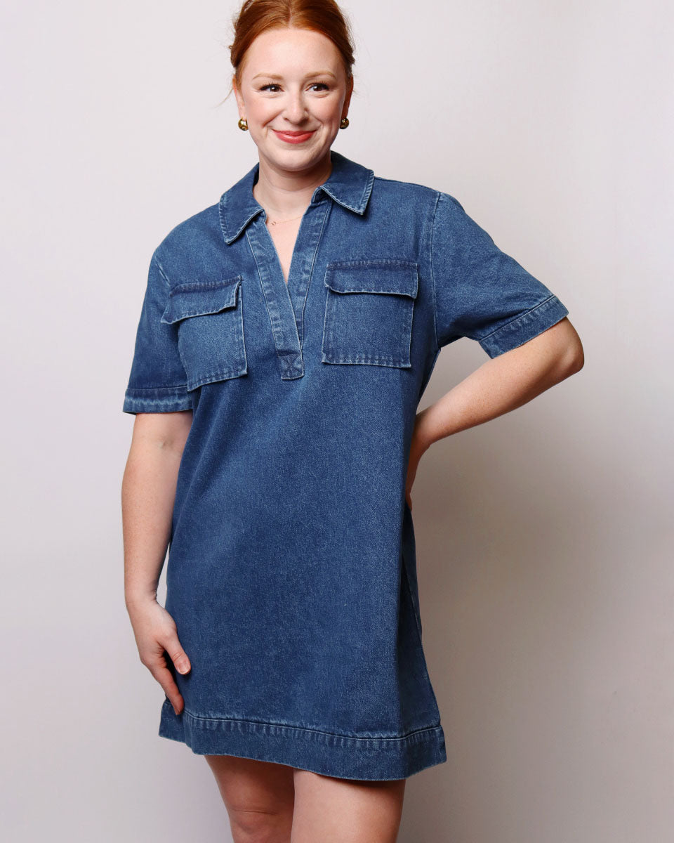A woman modeling a short-sleeve blue denim shirt dress against a plain light gray studio background.