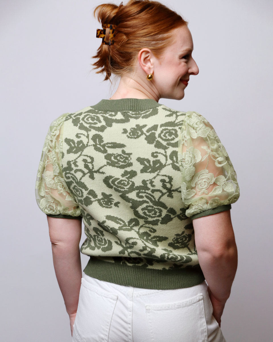 A woman with red hair tied up in a bun is shown from the back, wearing a sage green short-sleeve knit top with a darker green floral lace pattern and white pants, against a plain light gray background.