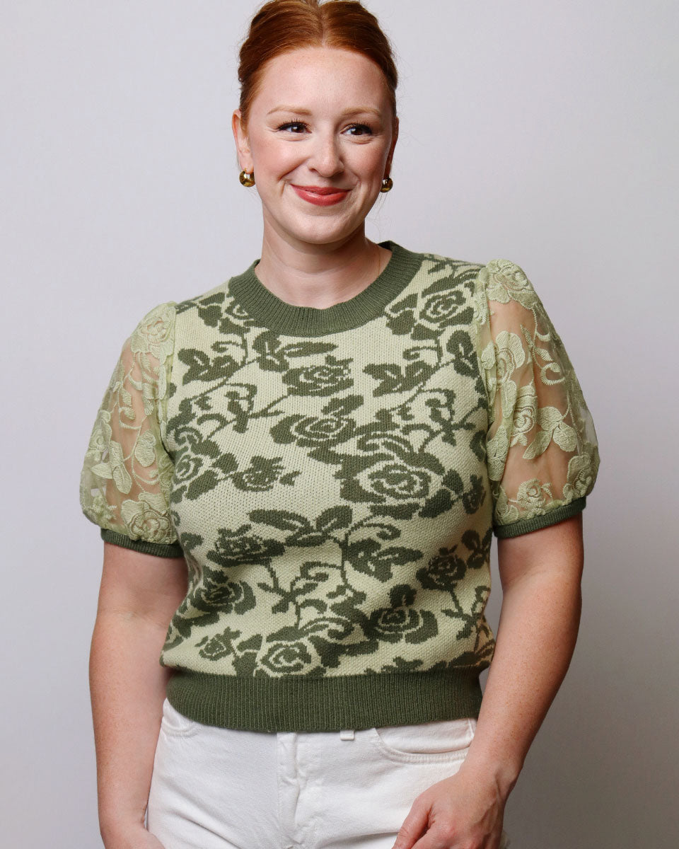 A woman with red hair tied up, wearing a sage green and olive green knit sweater with a floral rose pattern and sheer lace puff sleeves, paired with white pants, against a plain light gray background.