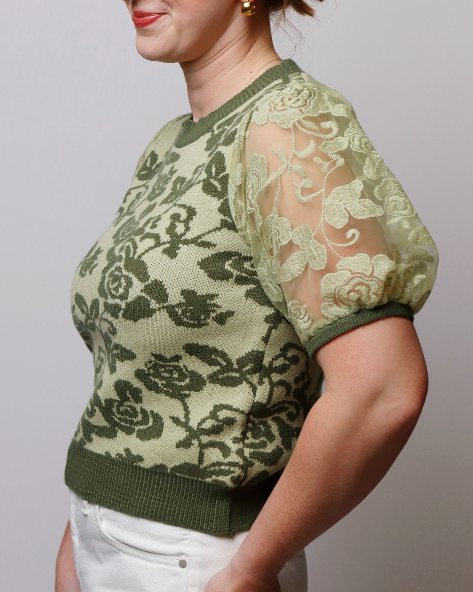 A woman wearing a cropped knit sweater with a sage green and olive green floral pattern, featuring sheer mesh puff sleeves with matching floral lace applique, paired with white pants.