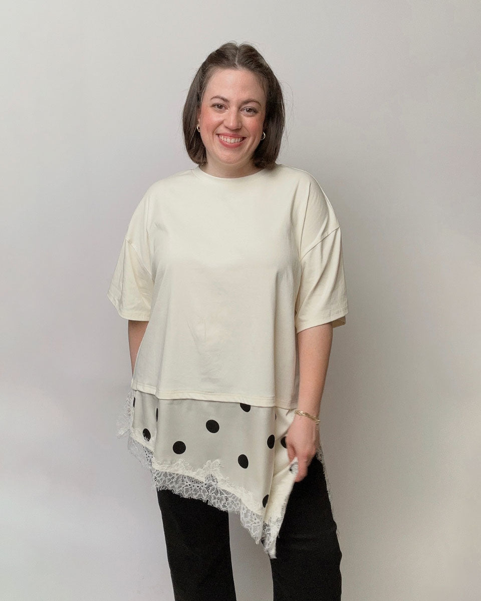 A woman modeling a cream-colored short-sleeve top with a sheer black polka-dot overlay and lace trim at the hem, paired with black pants, against a plain light gray background.