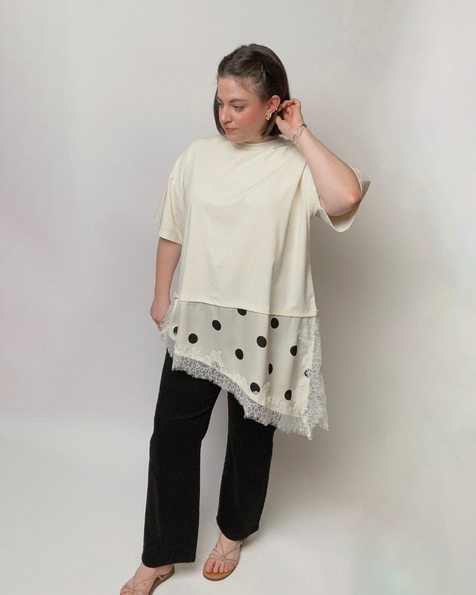 A woman modeling a cream-colored short-sleeve top with a sheer black and white polka dot lace hem, paired with black pants, against a plain light gray studio background.