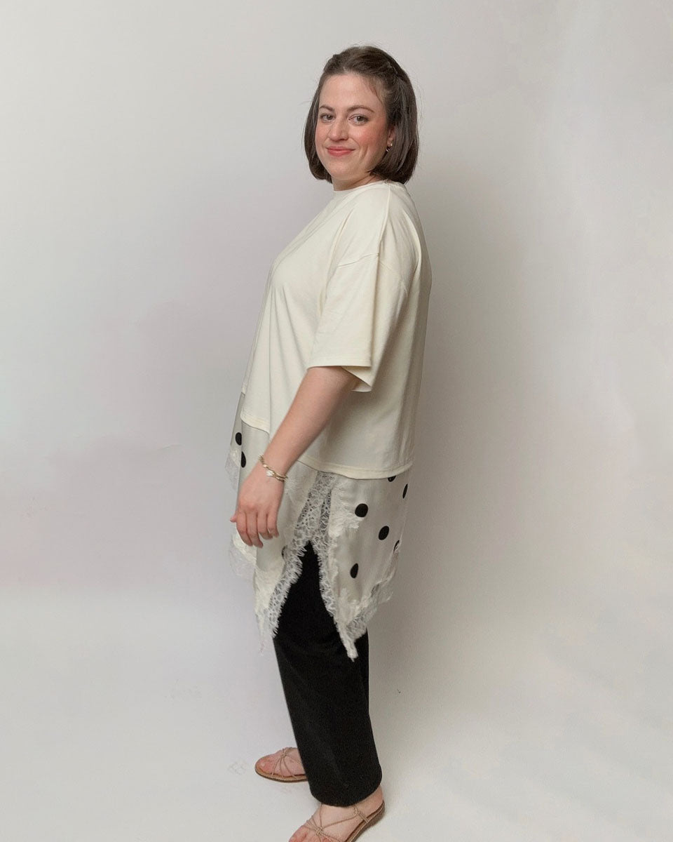 A woman modeling a cream-colored short-sleeve top with a sheer black polka-dot overlay and white lace trim, paired with black pants, standing against a light gray studio background.