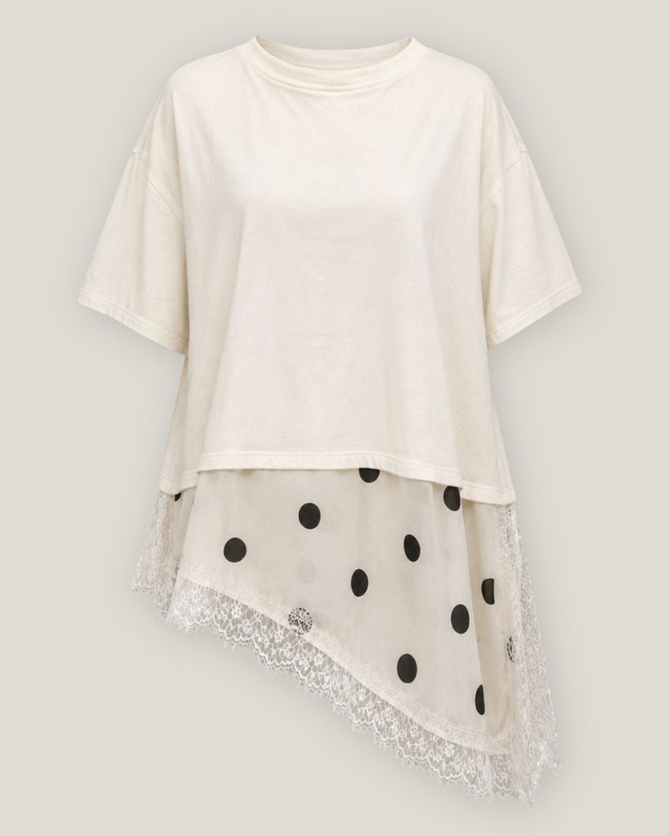 An asymmetrical cream-colored short-sleeve top featuring a sheer black polka-dot overlay and a white lace hem, displayed against a neutral light gray background.
