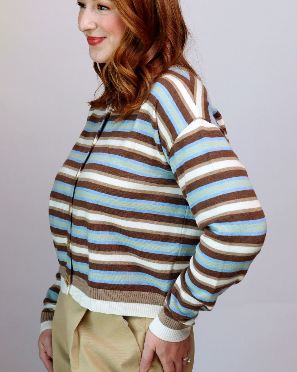 A woman wearing a cropped, long-sleeve knit cardigan with horizontal stripes in brown, light blue, cream, and white, paired with khaki pants, shown from a side profile against a light gray studio background.