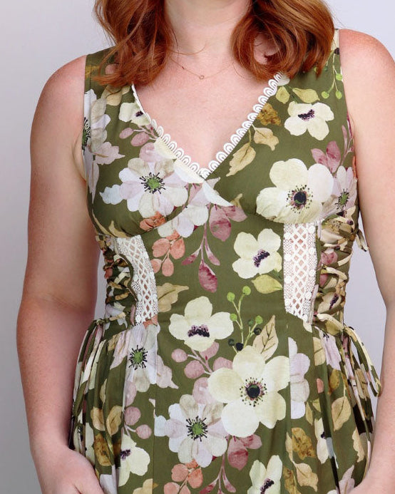 A woman with red hair wearing a sleeveless olive green floral dress with white and pink flowers, standing against a light gray studio background.