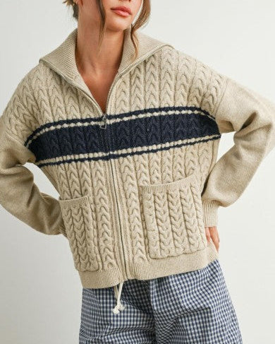 Woman wearing a beige cable knit sweater with a navy stripe on a plain background