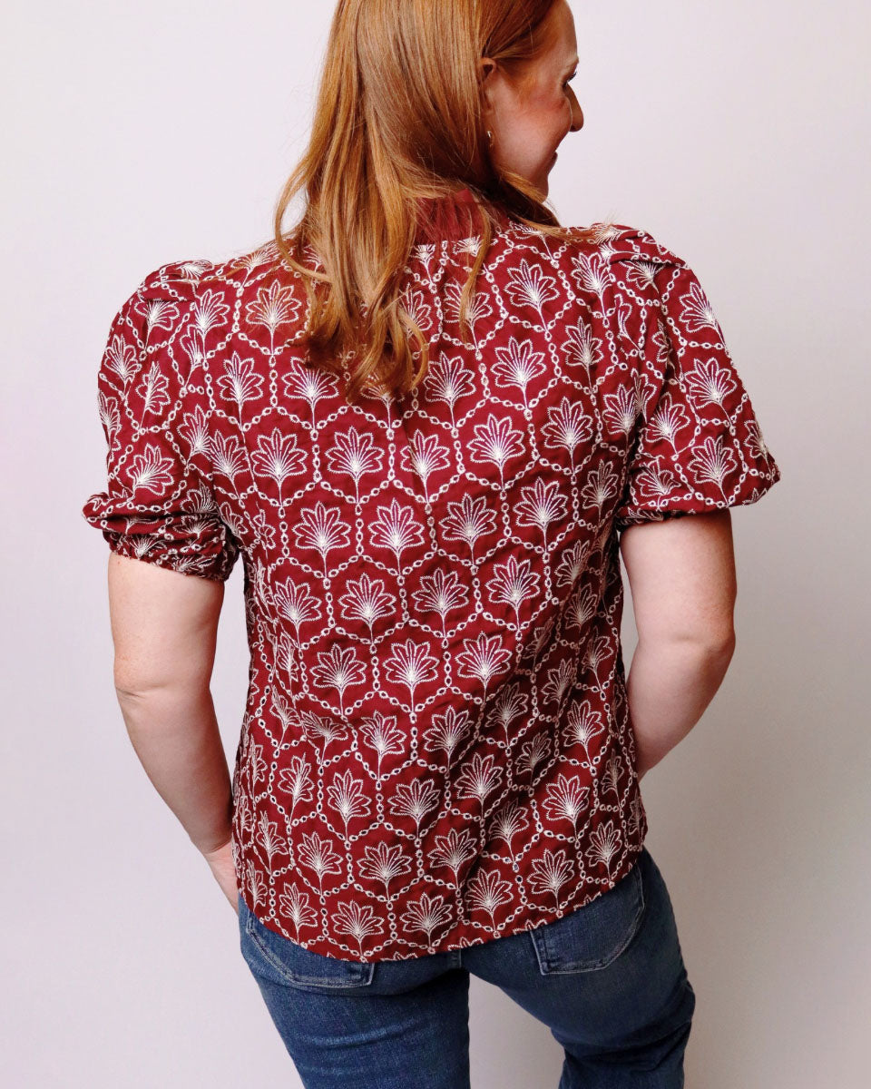 A woman viewed from behind wearing a short-sleeve button-up blouse with a red and white geometric pattern, paired with blue jeans, against a plain light gray background.