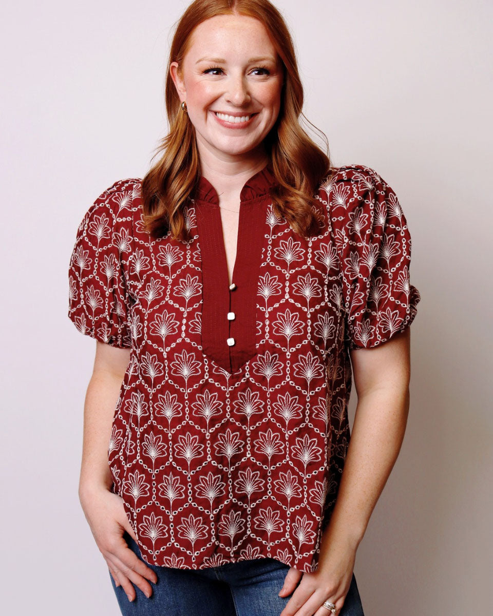 A smiling woman with red hair wearing a short-sleeve button-front blouse with puff sleeves. The blouse has a deep V-neckline with a small button placket and an allover white geometric and floral pattern on a dark red background.