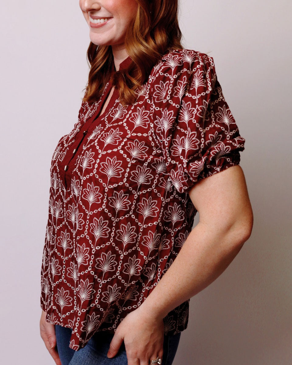 A woman wearing a short-sleeve blouse with a rust red and white floral pattern, paired with blue jeans, shown against a plain light gray background.