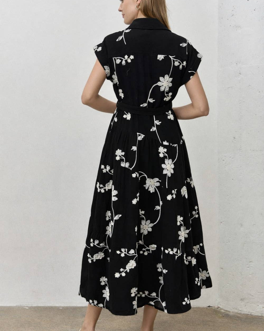 A woman wearing a black midi dress with a white floral pattern, shown from the back against a light gray textured wall.