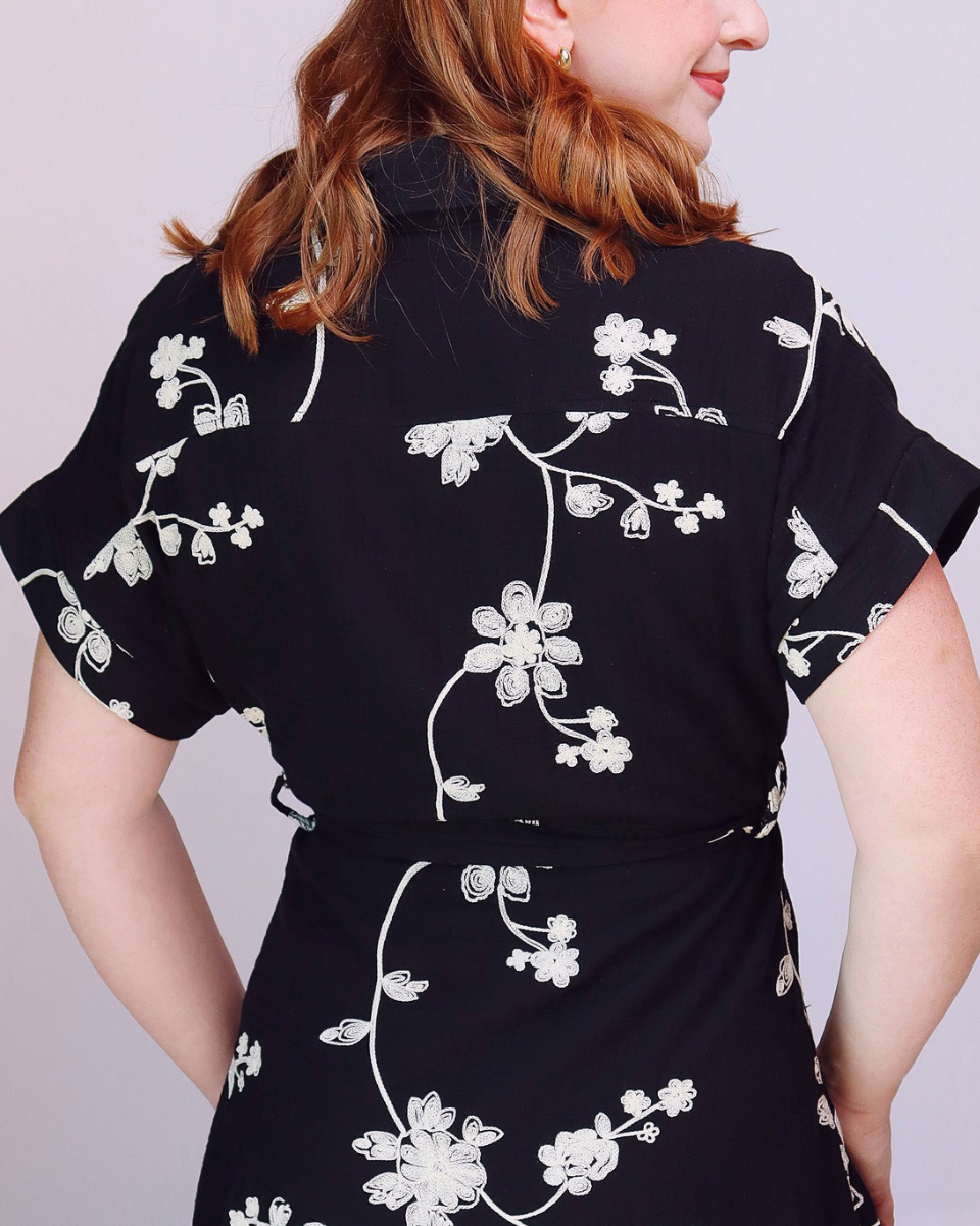 A woman shown from the back wearing a black short-sleeve dress with a white floral vine pattern, set against a plain light gray background.