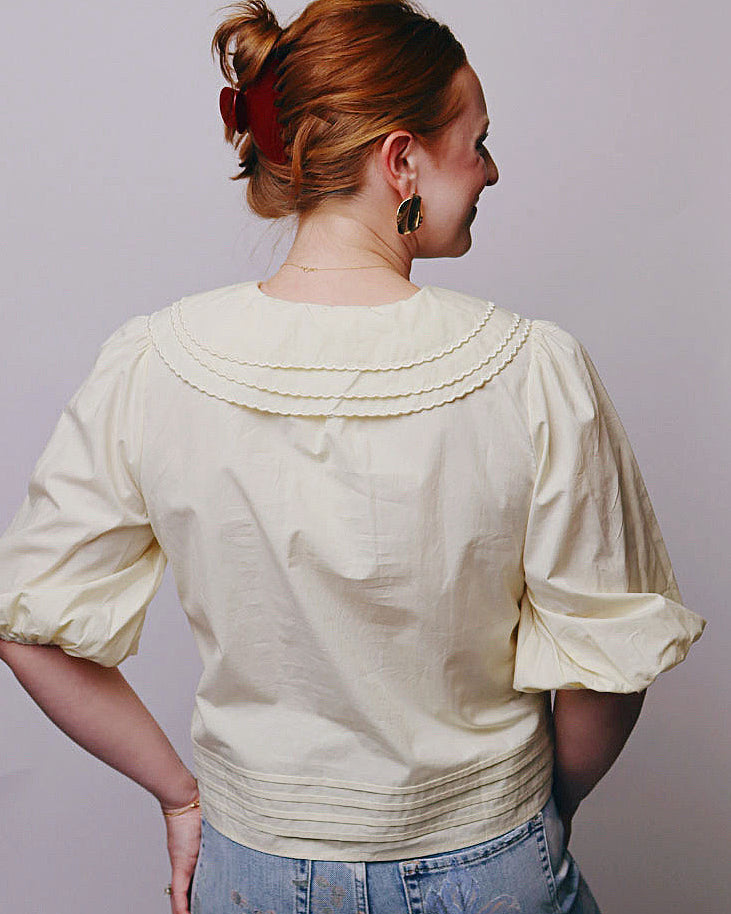 A woman shown from the back wearing a cream-colored blouse with a ruffled Peter Pan collar and puff sleeves, paired with light blue jeans, against a plain light gray background.