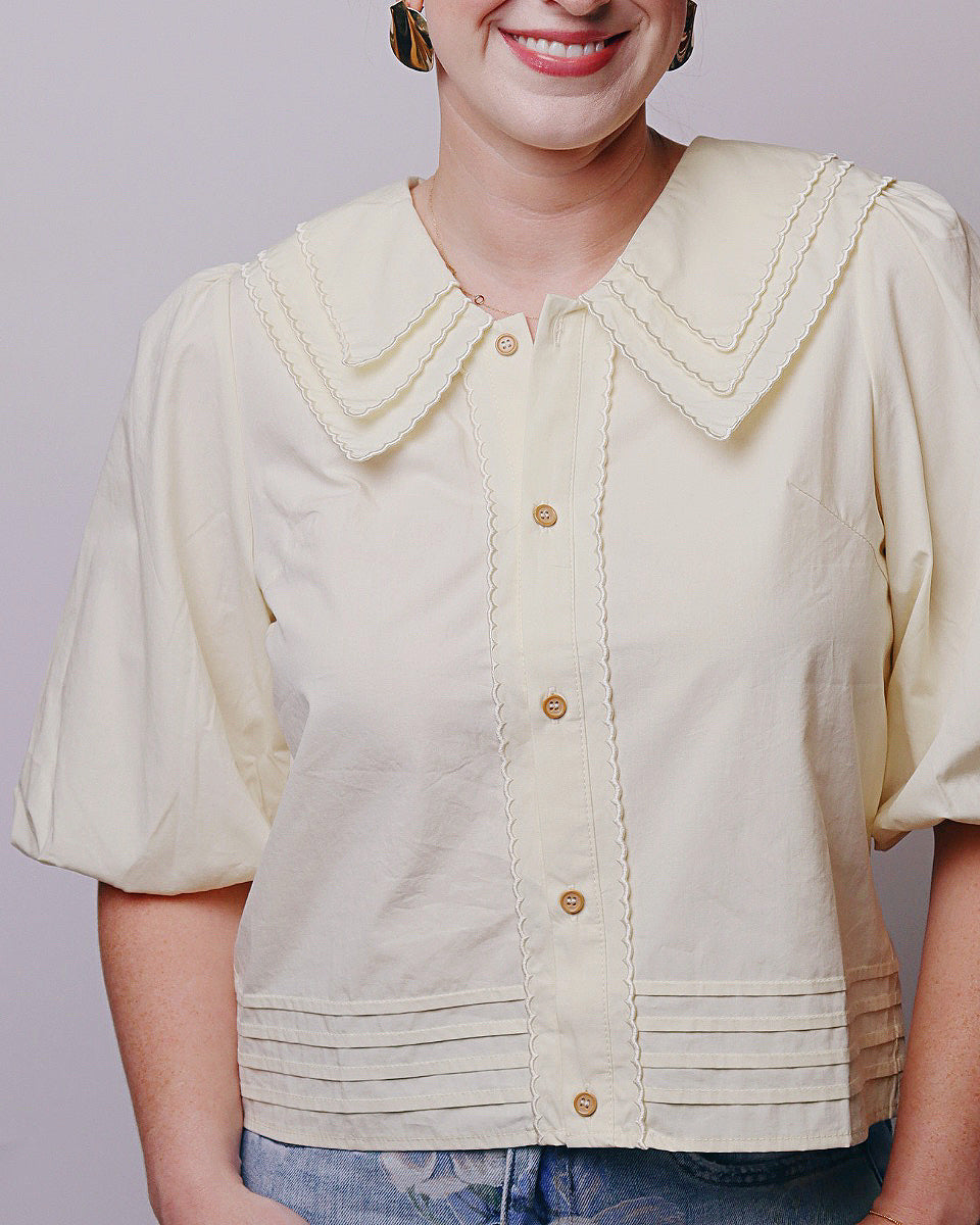 A woman wearing a cream-colored short-sleeve blouse with a large decorative collar, button-front closure, and gathered pleated detailing at the hem, paired with blue jeans against a plain light gray background.
