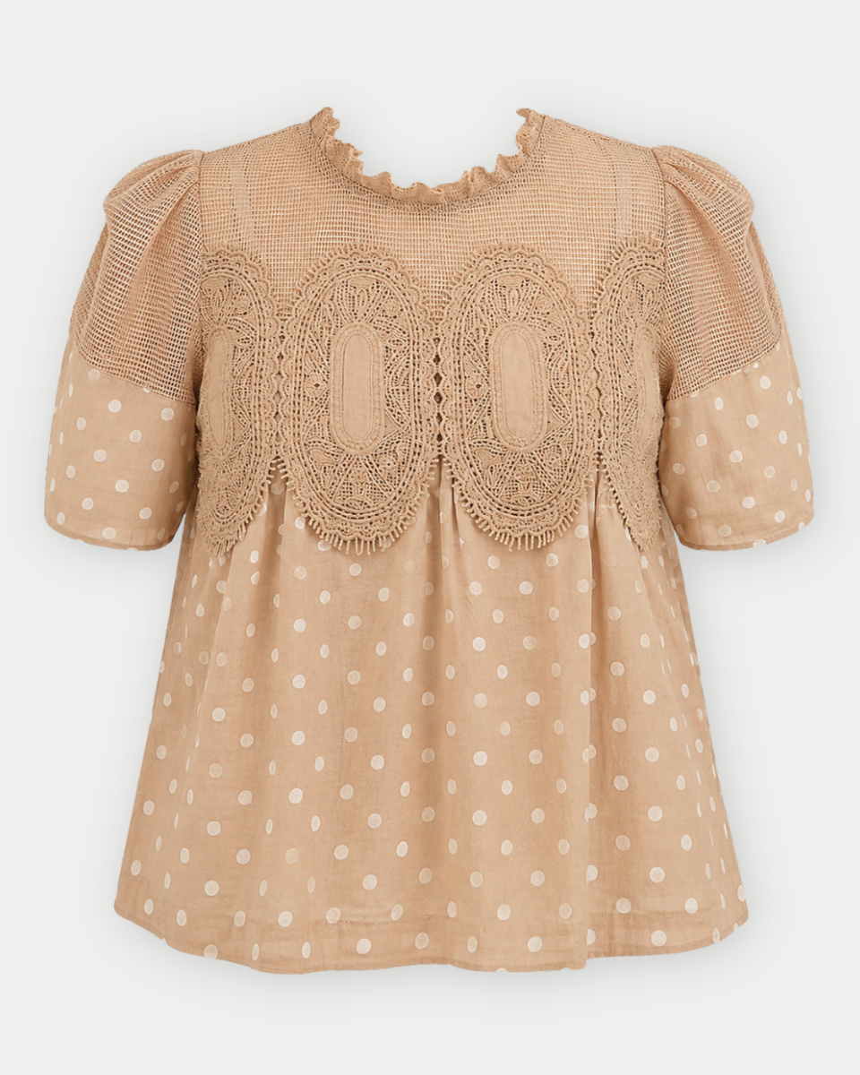 A beige short-sleeve blouse with white polka dots, featuring a ruffled round neckline and a decorative lace applique panel across the chest.