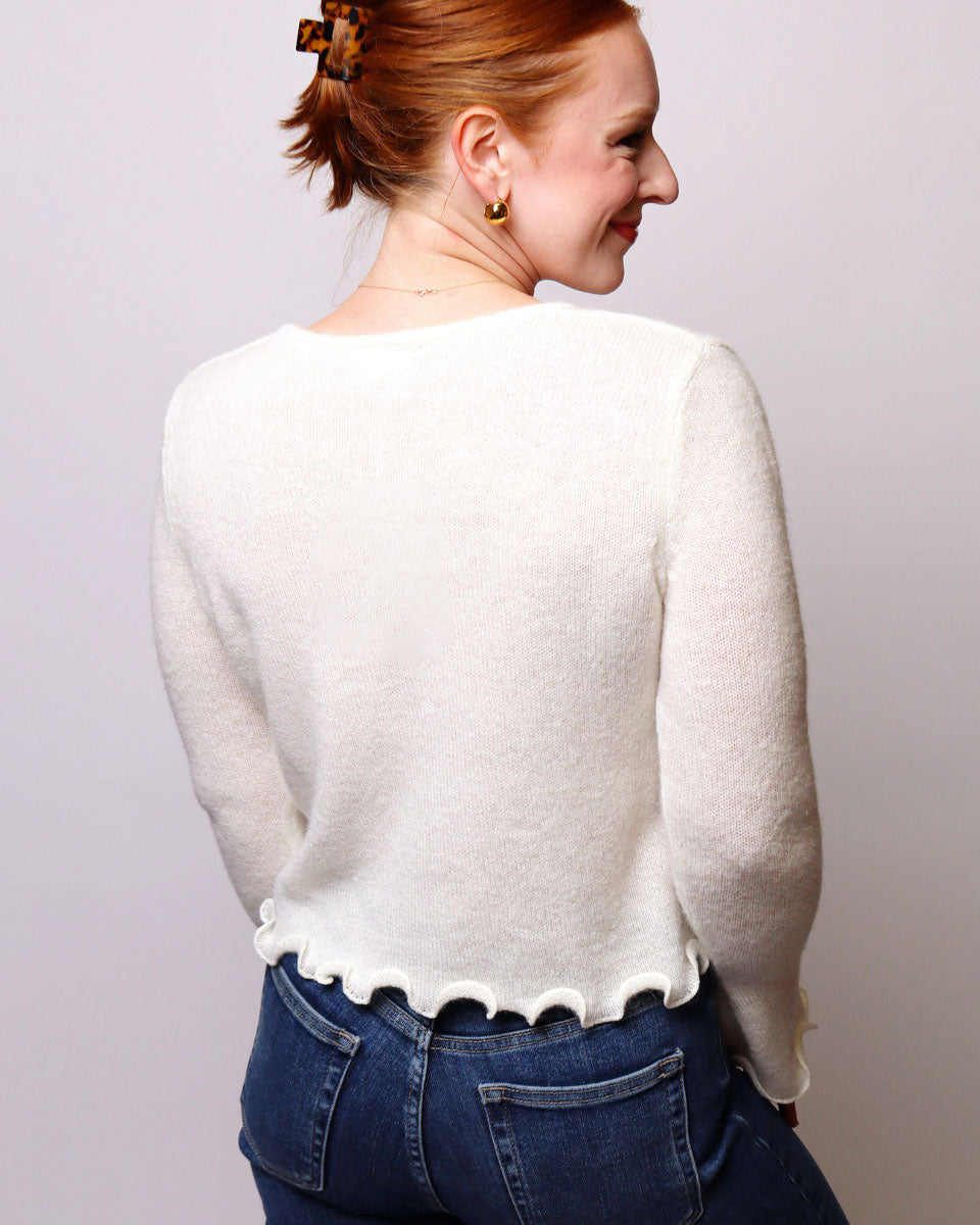 A woman with red hair tied up is shown from the back wearing a cream-colored, long-sleeve knit sweater with a scalloped hem and blue jeans, set against a plain light gray studio background.