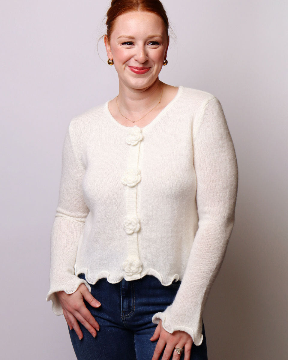 A woman wearing a cream-colored, long-sleeve knit cardigan with decorative flower buttons and a scalloped hem, paired with dark blue jeans, against a plain light gray background.