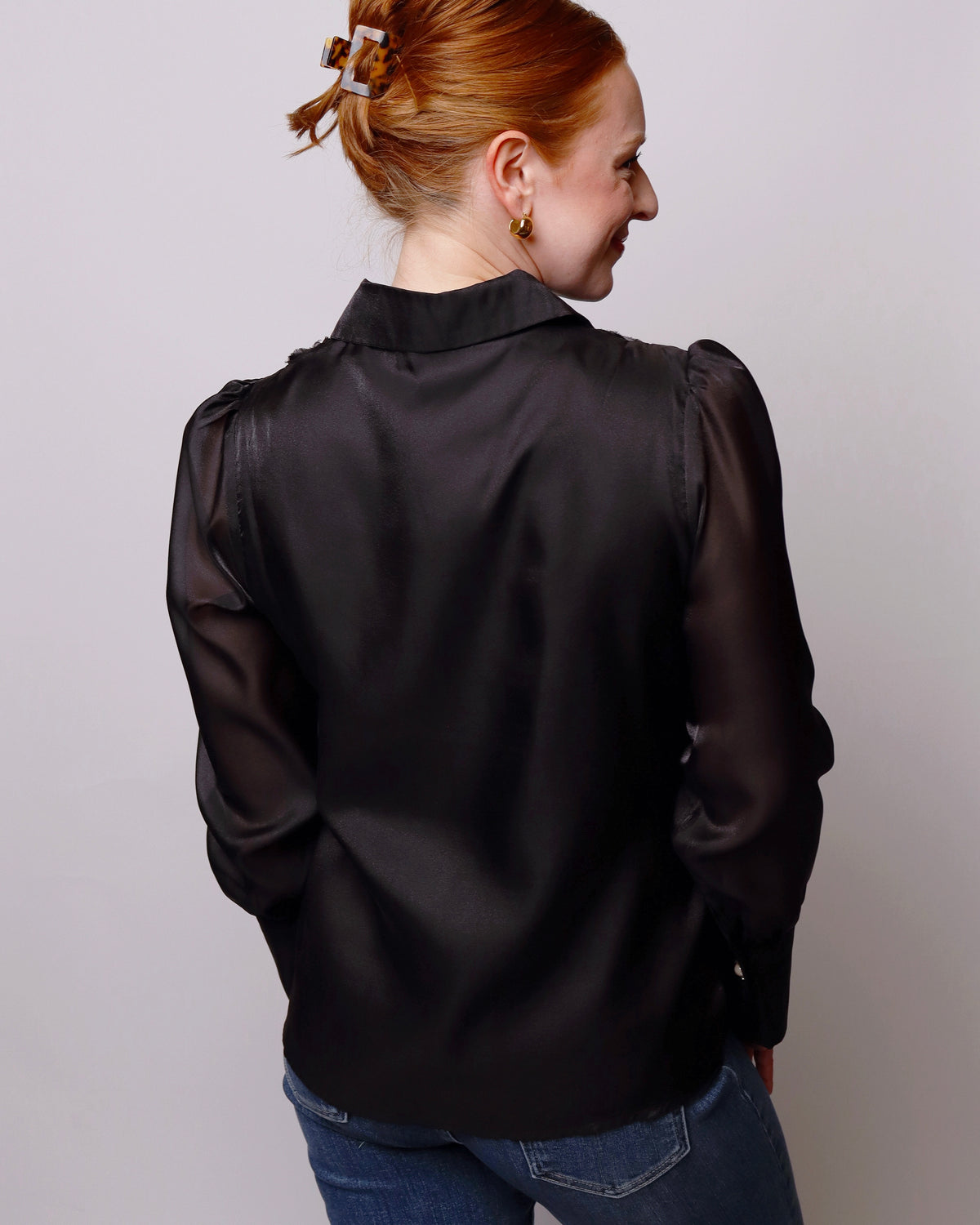 A woman with red hair tied up is shown from the back wearing a black satin blouse with sheer puff sleeves and blue jeans, set against a plain light gray background.