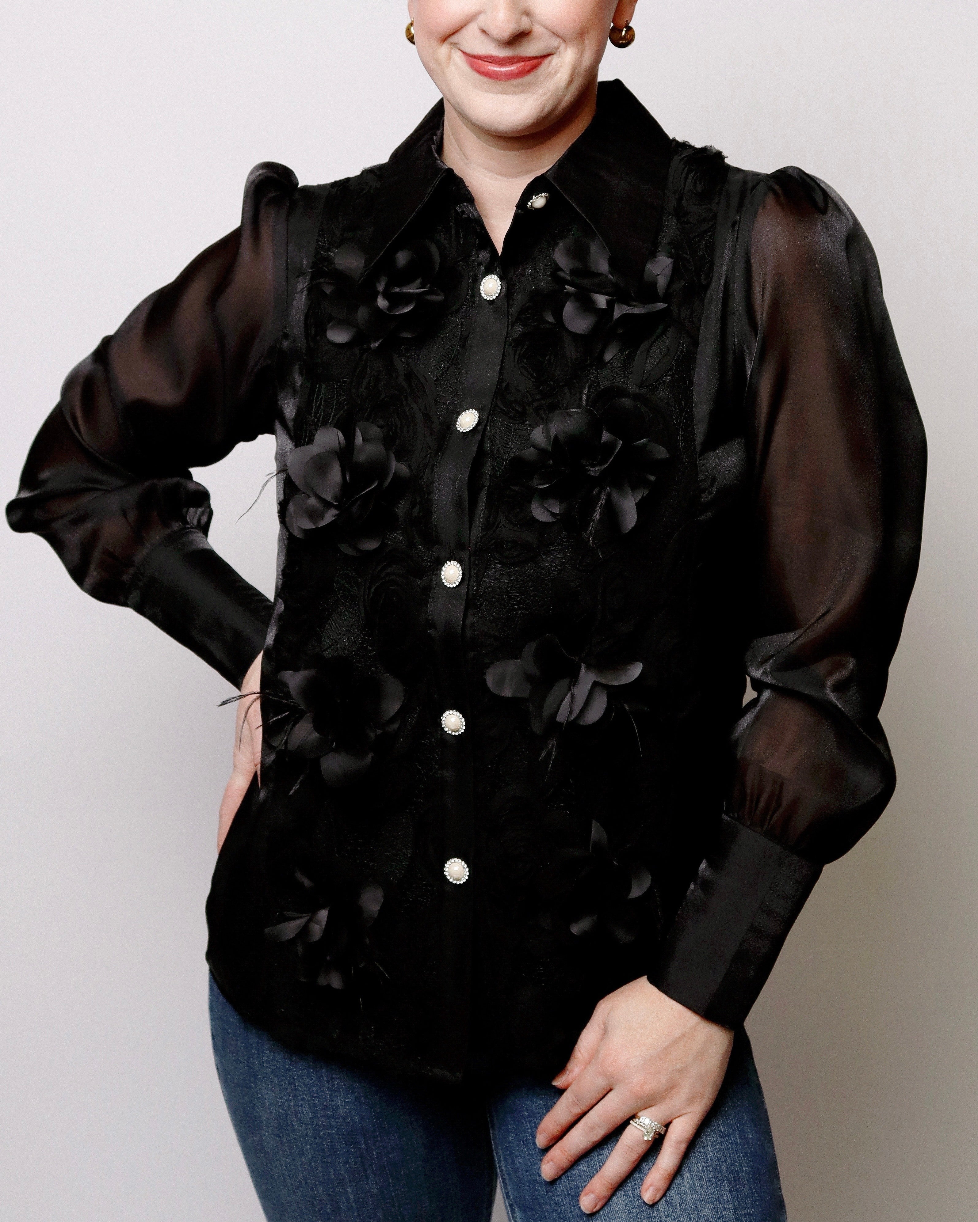 A woman wearing a black long-sleeve blouse with sheer puff sleeves and 3D floral applique, paired with blue jeans, against a light gray background.